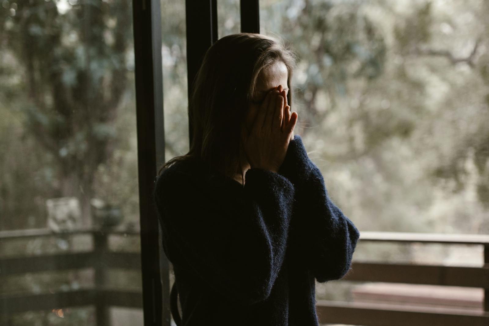 A contemplative woman stands by a window, hands over face, wearing a dark sweater.