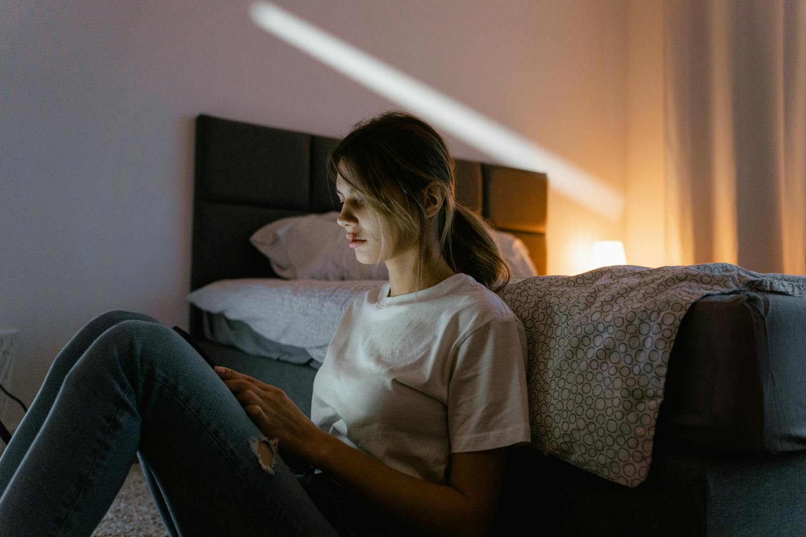 A young woman in a white shirt sits against a bed using a smartphone in a dimly lit bedroom.